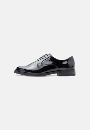 Chaussure habillée pour homme en cuir verni noir, avec laçage fermé et petit talon empilé, vue de profil sur fond blanc.