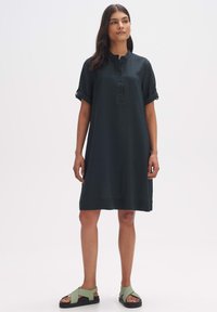 Dunkelgrünes Kurzarmkleid mit Knopfleiste, Stehkragen und umgekrempelten Ärmeln. Getragen mit hellgrünen Sandalen mit Kreuzriemen.