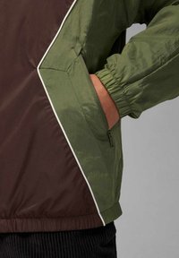 Väriblokkipusero tummanruskeassa ja oliivinvihreässä, jossa on kevyt materiaali, joustavat hihansuut ja vetoketjullinen taskudetalji.