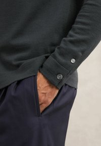 Dunkelgrauer Pullover aus strukturiertem Stoff, mit einem Bündchen und zwei runden Knöpfen, kombiniert mit navyblauen Hosen und einer sichtbaren Tasche.