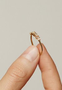 Anillo de oro con cinco piedras claras rectangulares y redondas, sostenido entre un pulgar y un dedo. Banda lisa con acabado pulido.