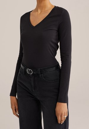 Persoon draagt een nauwsluitend zwart langarmshirt met V-hals, ingestopt in zwarte spijkerbroek met een zwarte riem met een zilveren gesp.