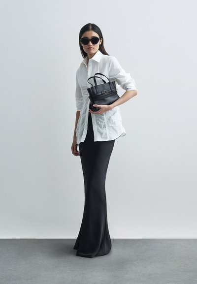 Γυναίκα με μαύρα γυαλιά ηλίου, λευκό πουκάμισο με πτυχές, μακριά μαύρη φούστα, κρατώντας μια μαύρη δερμάτινη τσάντα μπροστά σε απλό γκρίζο φόντο.