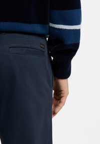 Marineblaue Hose mit glatter Textur, mit einer Gesäßtasche und einem kleinen, bestickten Logo. Kombiniert mit einem gestreiften Strickpullover in verschiedenen Blautönen.