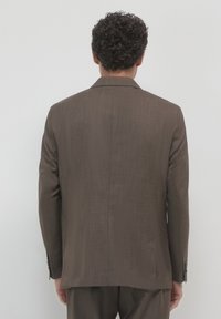Mann mit dunklem, lockigem Haar, der einen braunen Blazer und passende Hose trägt, mit dem Rücken zu einem neutralen, hellen Hintergrund gewandt.