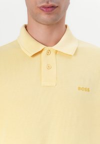 Polo-skjorte i lys gul bomull med krage, med to knapper og en gull "BOSS" logo på venstre bryst. Teksturert stoff.