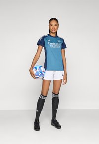 Kvinnors idrottsdräkt med en tealtröja med marinblå accenter, vita shorts, svarta strumpor med vita ränder och svarta fotbollsskor, som håller en färgglad fotboll.