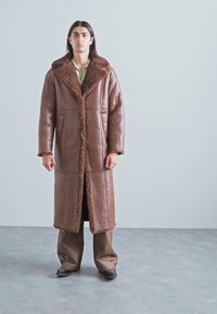 Uomo con lunghi capelli scuri che indossa un lungo cappotto di pelle scamosciata marrone, pantaloni larghi marroni e scarpe scure su un pavimento grigio con una parete chiara.