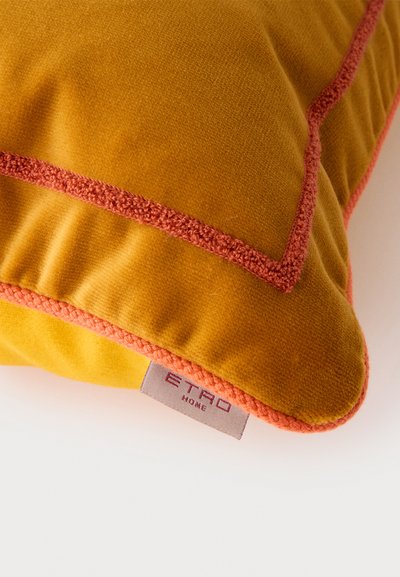 Coussin en velours jaune avec des bords passepoilés orange et une étiquette en tissu portant la mention "ETRO HOME" sur une surface blanche.