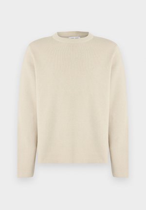 Pull beige à manches longues avec col rond et un petit logo subtil sur la poitrine gauche, présenté sur un fond blanc uni.