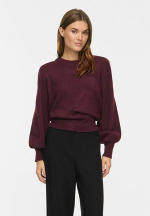 Femme portant un pull bordeaux à manches longues avec des manches bouffantes et un pantalon noir taille haute, debout devant un fond blanc uni.
