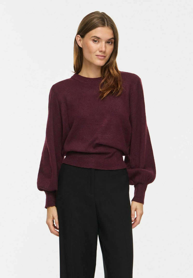 Femme portant un pull bordeaux à manches longues avec des manches bouffantes et un pantalon noir taille haute, debout devant un fond blanc uni.