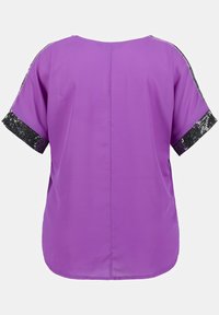 Lila blus med korta ärmar, prydd med svarta paljettdetaljer på ärmsluten. Slät tyg med rak skärning och rundad nederkant.