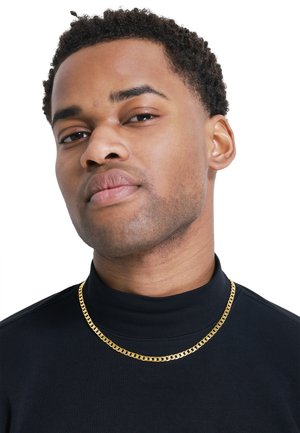 Gullkjede på en modell som har på seg en svart turtleneck. Kjedet har en flat, sammenflettet design som gir glans mot stoffet.