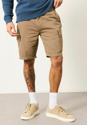 Mann trägt khakifarbene Cargo-Shorts, beige Sneakers, weiße Socken und ein blaues langärmliges Hemd und steht mit einer Hand in der Nähe der Tasche.
