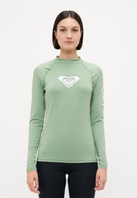 Lys grønn langermet rash guard med høy hals. Har en hvit hjerte-logo på brystet og fet tekst på ermet. Glatt tekstur.