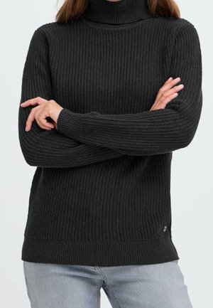 Personne portant un pull à col roulé noir côtelé à manches longues, les bras croisés, et un pantalon gris clair sur un fond uni.