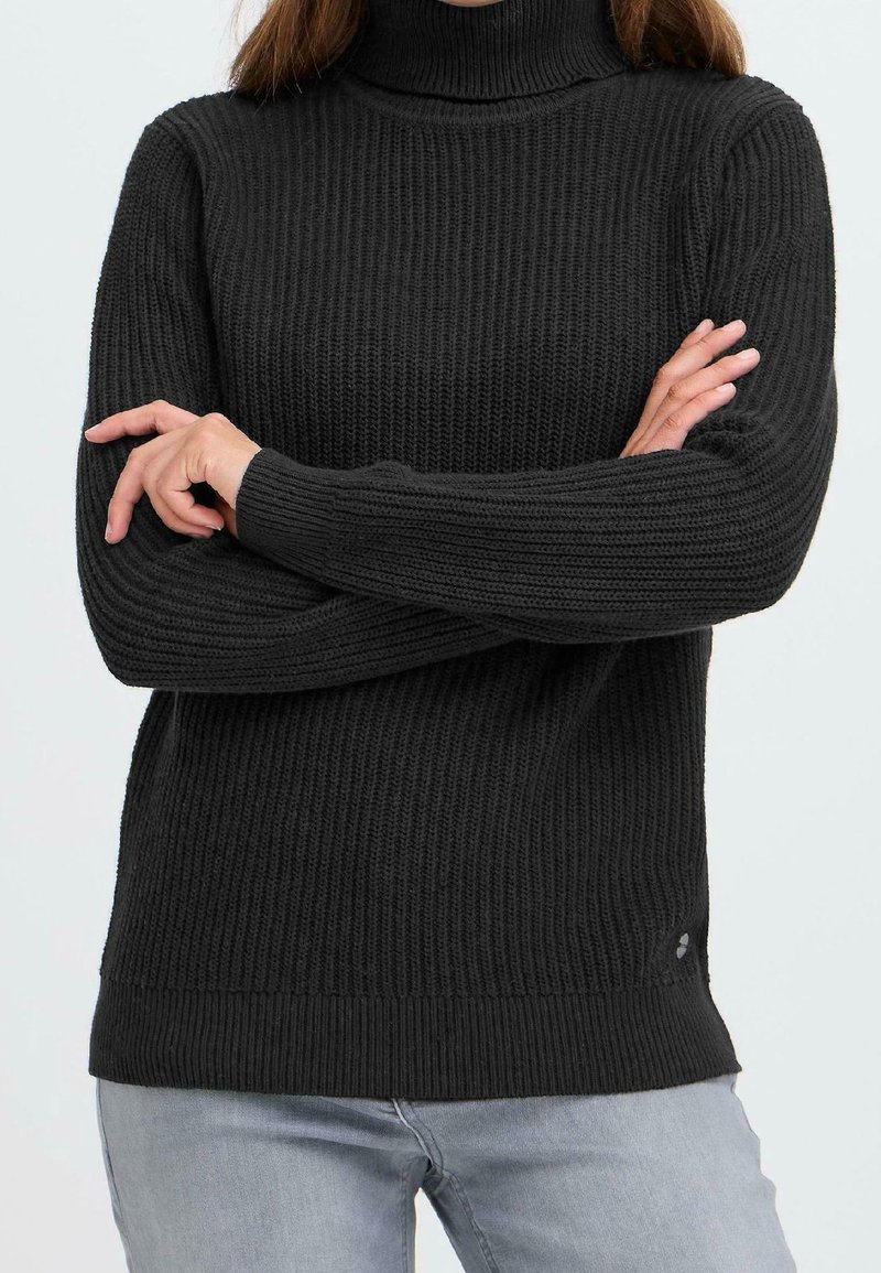 Personne portant un pull à col roulé noir côtelé à manches longues, les bras croisés, et un pantalon gris clair sur un fond uni.