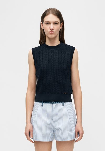 Γυναίκα που φοράει αμάνικο μπλε σκούρο πλεκτό τοπ με σχέδιο πλεξούδα και υψηλόμεσο σορτς με κάθετες ριγέ σε ανοιχτό μπλε και λευκό, στέκεται μπροστά σε λευκό φόντο.
