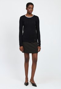 Suéter negro de punto con un patrón de trenzado, cuello redondo, mangas largas, combinado con una mini falda negra y zapatos de punta negros.