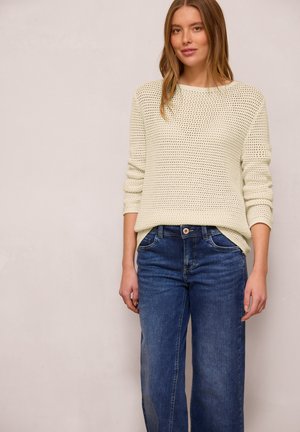 Žena stojí oblečená v krémovém pleteném svetru s vyhrnutými rukávy a modrých rovně střižených džínách na jednobarevném světlém pozadí.