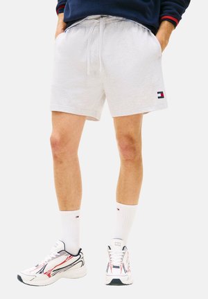 Hombre con pantalones cortos de cordón gris claro con bolsillos, calcetas blancas hasta el tobillo y zapatillas blancas con detalles en rojo y negro, de pie con las manos en los bolsillos.