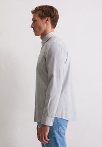 Szara koszula w paski na guziki z kołnierzykiem, długimi rękawami i widocznymi guzikami. Nosi się ją z jasnoniebieskimi dżinsami, pokazana z boku.