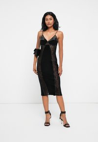 Μαύρο φόρεμα με δαντελένιες λεπτομέρειες, εφαρμοστή σιλουέτα και λεπτές τιράντες. Φοριέται με μαύρες ψηλές γόβες και μια μικρή μαύρη τσάντα.