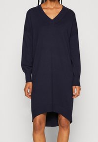 Robe pull bleu marine à manches longues, décolleté en V et manches côtelées. Présente un ourlet arrière légèrement plus long et une texture douce.
