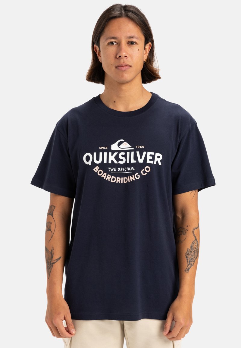 Camiseta Quiksilver azul marino con cuello redondo. Presenta un logo blanco centrado en el pecho, mangas cortas y un ajuste relajado.