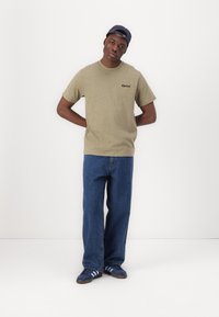 T-shirt in cotone verde oliva con logo, abbinata a jeans denim azzurri larghi e sneakers navy con strisce azzurre.