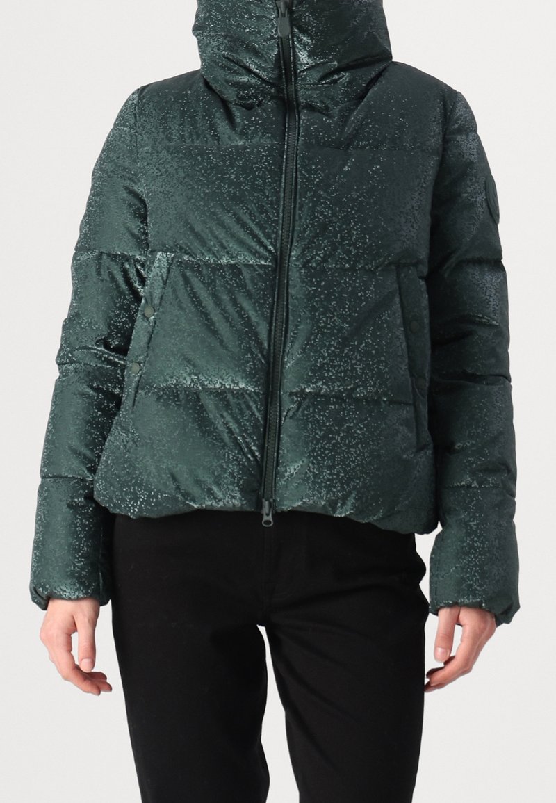 Doudoune verte foncée tachetée avec fermeture éclair à l'avant et poches à boutons-pression, portée avec un pantalon noir, vue du cou aux hanches.