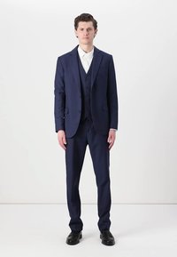 Traje azul marino con chaqueta ajustada y pantalones a juego. Tejido texturizado, chaleco de cinco botones debajo de la chaqueta, combinado con una camisa blanca.