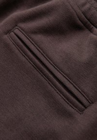 Gros plan d'un tissu texturé brun foncé avec une poche passepoil étroite et des coutures visibles le long des bords.