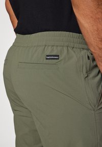 Oliven grønne træningsbukser med elastisk talje, der har en baglomme og et lille sort logo-patch bagpå. Glat stofoverflade.