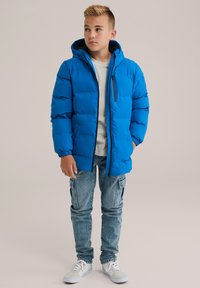 Chaqueta acolchada azul con cierre frontal y capucha, con puños elásticos. Lleva puesta sobre una camisa ligera y combinado con vaqueros de mezclilla claros y zapatillas deportivas.