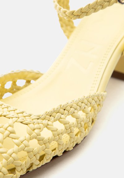 Κίτρινο υφασμάτινο δερμάτινο σανδάλι με πλεκτά λουριά και τετράγωνο τακούνι, εμφανισμένο σε κοντινό πλάνο σε λευκό φόντο.