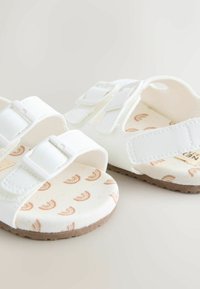 White sandals featuring two adjustable straps, a soft, patterned insole with rainbow motifs, and a textured brown outsole.
