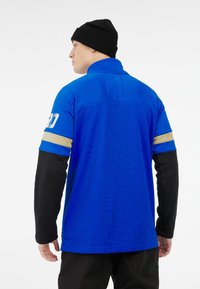 Blauwe sportieve pullover met een mesh-textuur. Voorzien van een hoge kraag en zwarte mouwen, geaccentueerd met een gouden streep op de mouw.