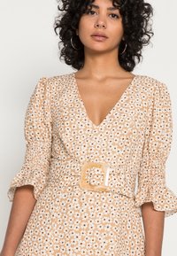 Vestido com padrão floral em bege, com mangas curtas em balão e um decote em V profundo, apresentando uma fivela de cinto quadrada texturizada e uma cintura franzida.