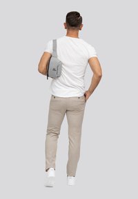 Sac à épaule gris en matériau lisse, de forme rectangulaire, arborant un logo. Porté par-dessus une chemise blanche et un pantalon beige, avec un design minimaliste.