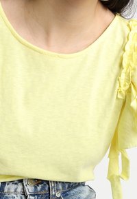Žlutý bavlněný top s krátkými volánkovými rukávy, s výstřihem ve tvaru lodičky a měkkou texturou. V kombinaci s džínovými šortkami.