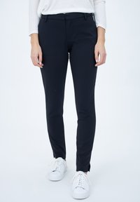 Pantaloni neri aderenti con una texture liscia, tasche frontali e una vita a media altezza, abbinati a sneaker bianche.