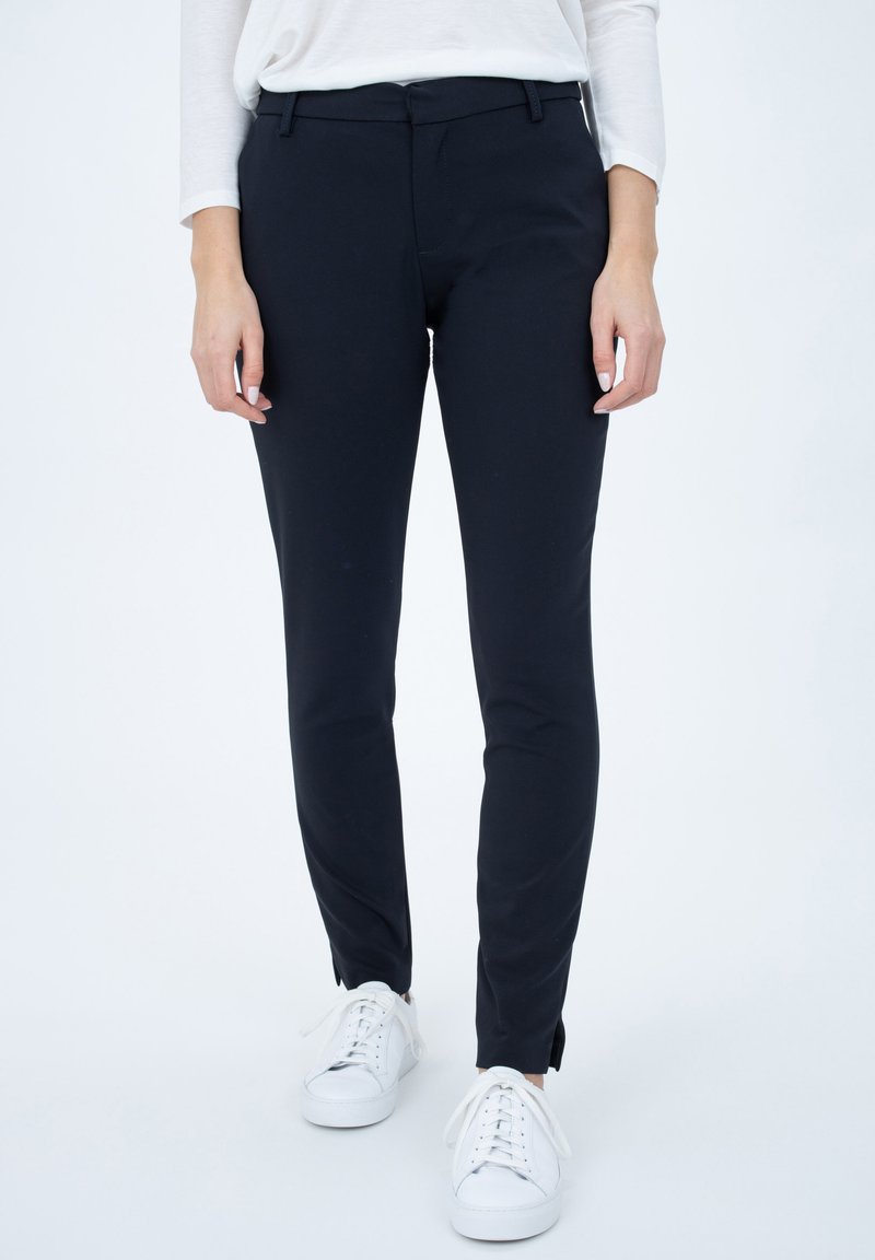 Pantaloni neri aderenti con una texture liscia, tasche frontali e una vita a media altezza, abbinati a sneaker bianche.