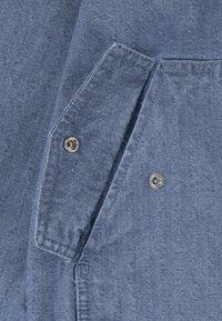 Näruppfattning av blå denimtyg som visar en ficka med tryckknapp och synliga sömnadsdetaljer. Texturen ser mjuk och använd ut.
