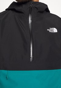 Giacca a vento nera e teal con zip frontale, dotata di cappuccio e del logo North Face in bianco sul lato sinistro del petto. Tessuto liscio.