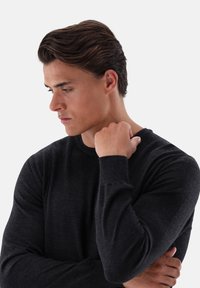 Schwarzer Strickpullover mit Rundhalsausschnitt, gerippten Bündchen und strukturierter Oberfläche. Modell posiert mit verschränkten Armen vor einem neutralen Hintergrund.