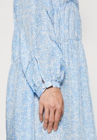 Vestido floral de color azul claro con una textura suave, mangas recogidas y detalles de puños abotonados. Presenta un delicado estampado de pequeñas flores blancas.