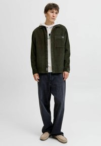 Donkergroene corduroy overhemdjasje met een voorzak, gedragen over een witte hoodie, gecombineerd met losse donkere spijkerbroek en beige schoenen.