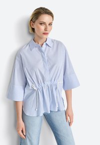 Blouse rayée bleu et blanc avec taille à cordon, manches surdimensionnées et col à boutons, en tissu léger.